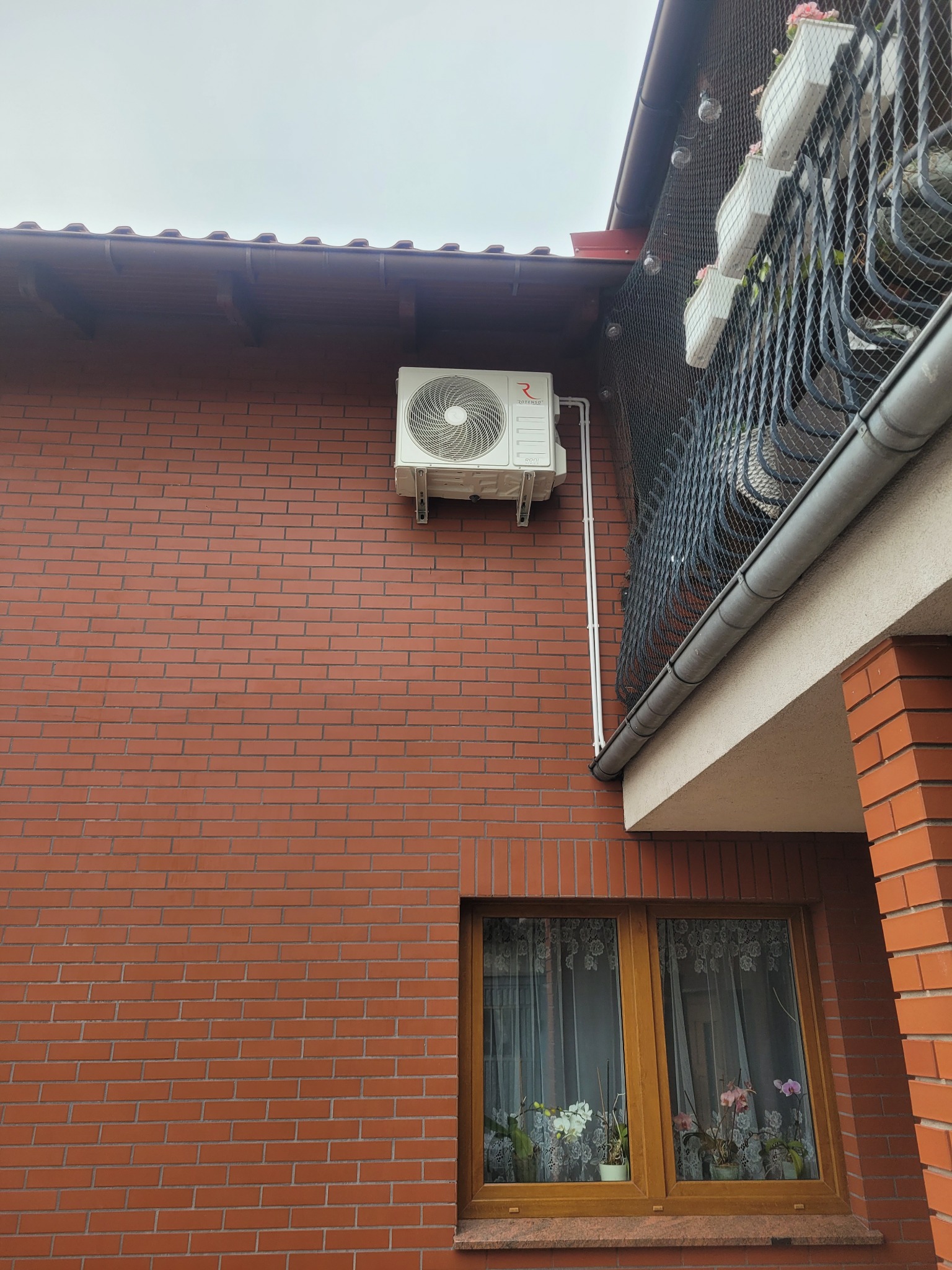 Biały klimatyzator Rotenso zamontowany na ceglanej ścianie domu, z widocznymi rurami odprowadzającymi. Na balkonie siatka zabezpieczająca i kwiaty.