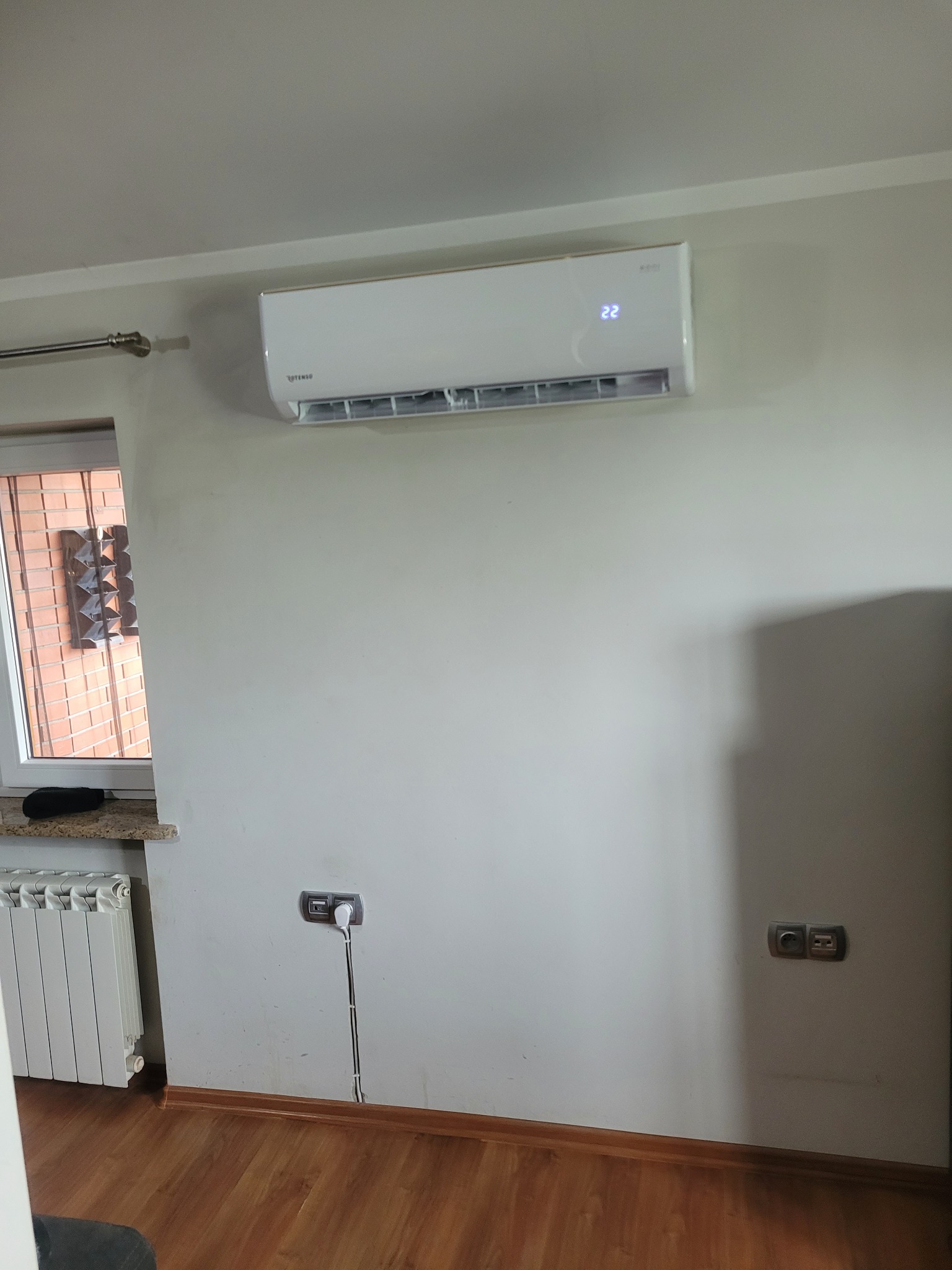 Biały klimatyzator Rotenso z wyświetlaczem temperatury (22°C) zamontowany na ścianie w jasnym wnętrzu, widoczne gniazdka elektryczne i fragment grzejnika.