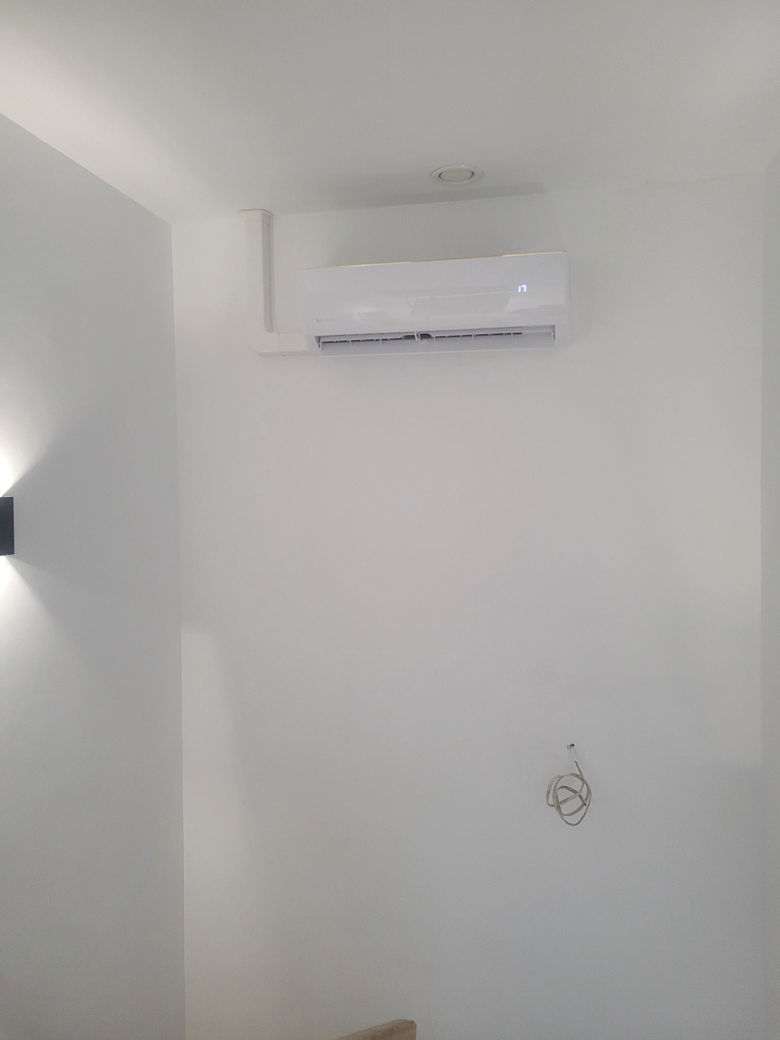 Biały klimatyzator ścienny z wyświetlaczem temperatury (17 stopni Celsjusza) zamontowany w minimalistycznym wnętrzu z widocznymi przewodami.