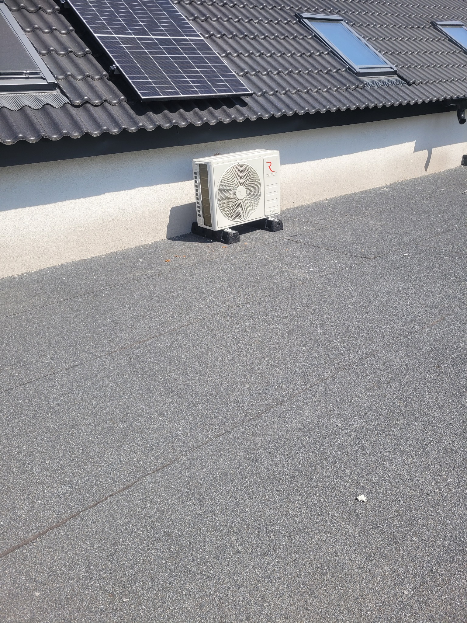 Biały klimatyzator Rotenso NOORI na dachu z panelami słonecznymi i oknami dachowymi w tle, widok z góry.