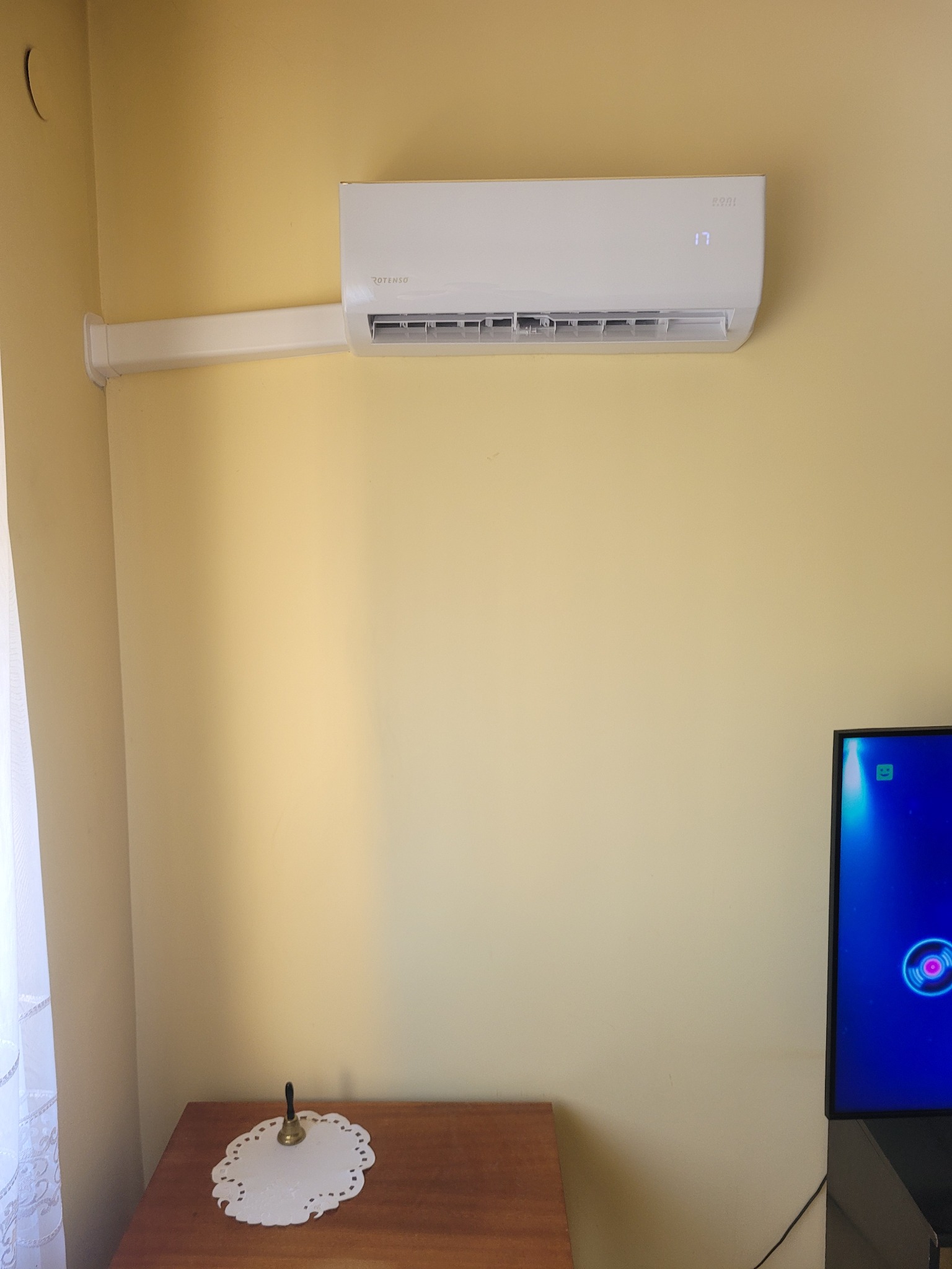 Biały klimatyzator Rotenso z długą listwą maskującą przewody zamontowany na żółtej ścianie w pokoju. Na urządzeniu wyświetla się temperatura 17 stopni.