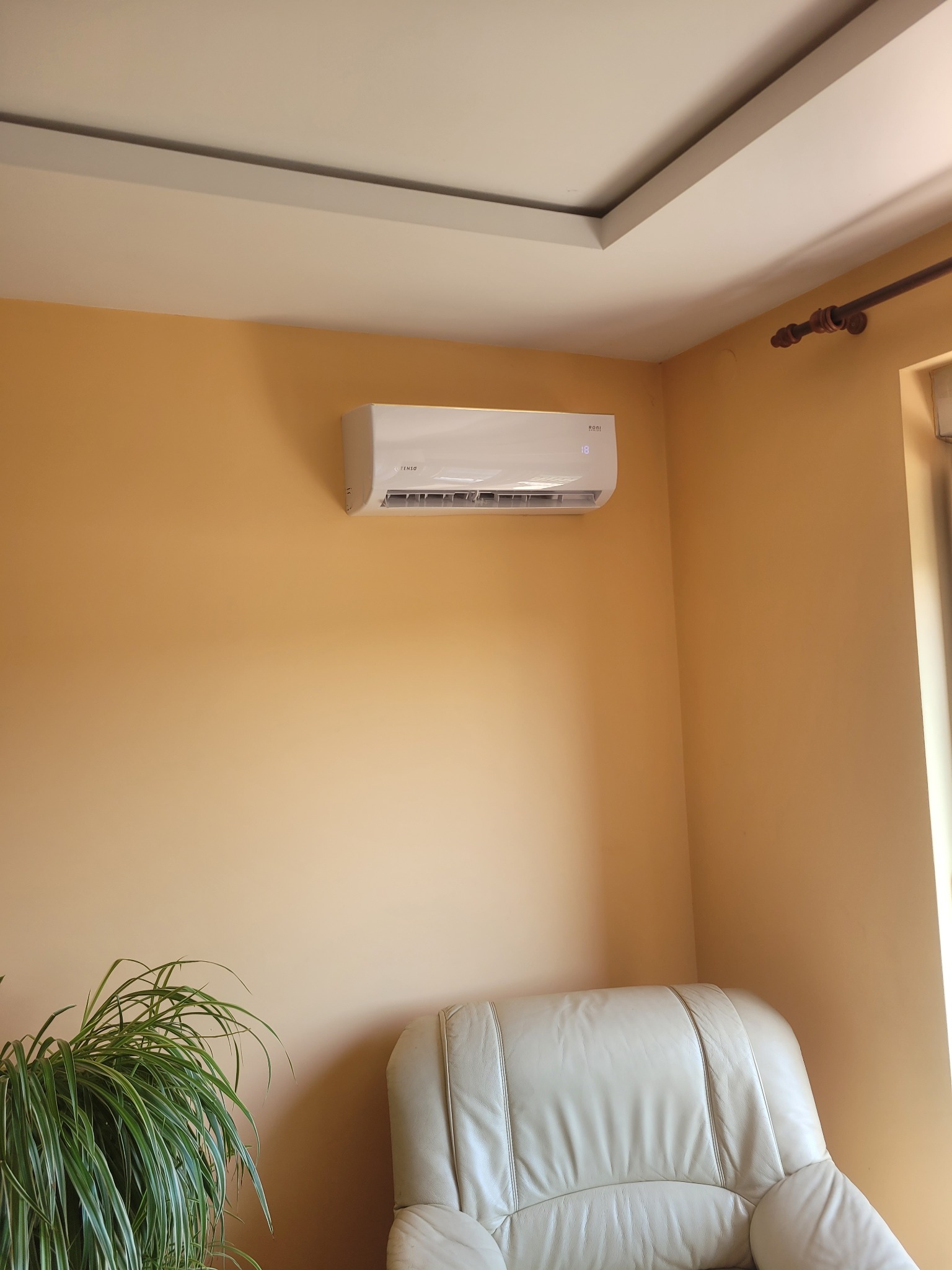 Klimatyzator ścienny marki Fenso zamontowany w pokoju z beżowymi ścianami. Widoczny fragment sofy i doniczka z zieloną rośliną. Na wyświetlaczu klimatyzatora widoczna temperatura 18 stopni.