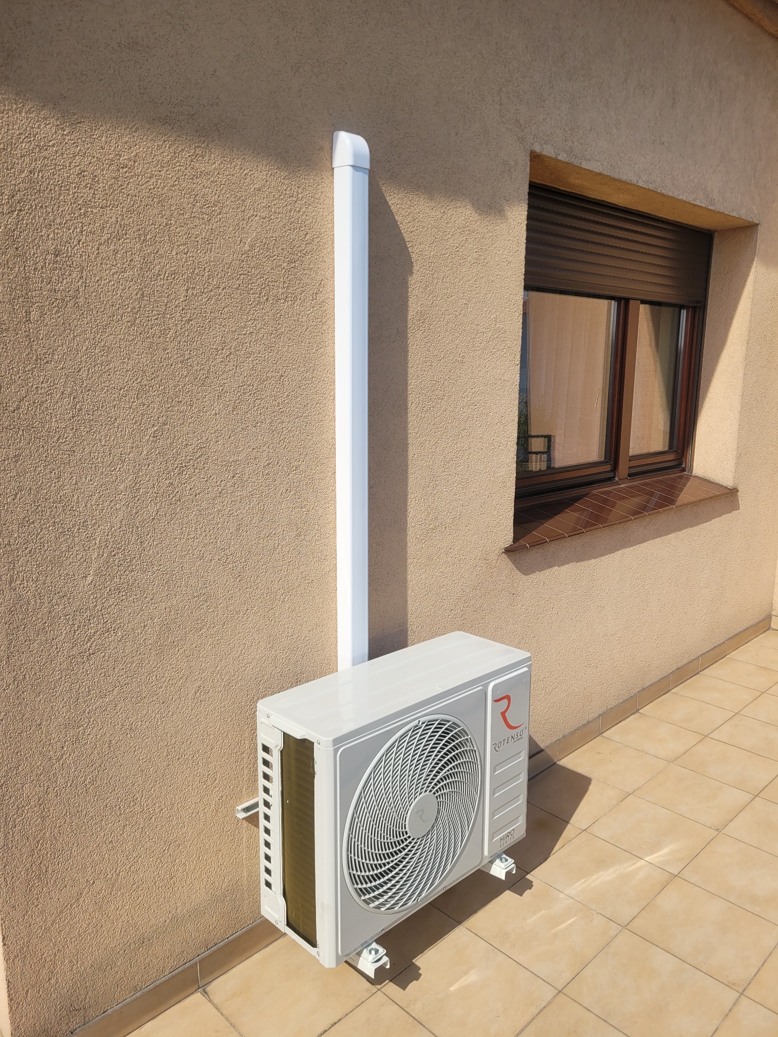 Zamontowana, biała jednostka zewnętrzna klimatyzacji Rotenso Hiro Series na elewacji budynku z oknem z brązowymi roletami zewnętrznymi i białym odprowadzeniem skroplin.