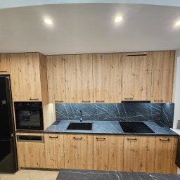 hm-meble marcin heinrich - Kompletna zabudowa kuchenna z drewnopodobnymi frontami, czarnym blatem z marmurkowym wzorem i czarnym sprzętem AGD, obejmująca szafki górne i dolne, piekarnik, płytę grzewczą i zlew, oświetlona...