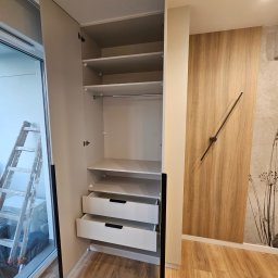 hm-meble marcin heinrich - Jasna szafa z otwartymi drzwiami i wysuniętymi szufladami, widoczna drabina oparta o okno balkonowe, drewniana okładzina ścienna z minimalistycznym zegarem.