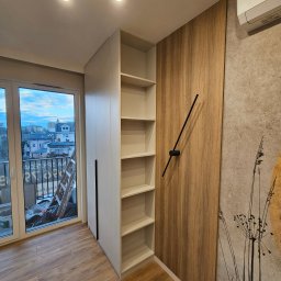 hm-meble marcin heinrich - Bydgoszcz: Biała szafa wnękowa z półkami sąsiadująca z drewnianą ścianą z minimalistycznym zegarem, widok z okna na miasto.