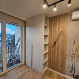 hm-meble marcin heinrich - Jasnoszara szafa wnękowa z czarnymi uchwytami, z otwartymi półkami, na podłodze panele, na ścianie dekoracyjny panel drewnopodobny z zegarem, widok na balkon z drabiną