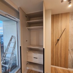 hm-meble marcin heinrich - Otwarta, jasna szafa wnękowa z półkami i szufladą, widoczna drabina oparta o okno balkonowe, drewniana okładzina ściany z minimalistycznym zegarem.