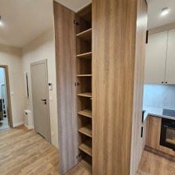 hm-meble marcin heinrich - Wysoka, drewniana szafa wnękowa z otwartymi drzwiami, ukazująca puste półki, wkomponowana w przestrzeń między przedpokojem a kuchnią, z podłogą imitującą drewno.