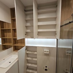 hm-meble marcin heinrich - Nowoczesna łazienka z szafkami z białymi frontami i drewnianymi półkami, otwarte drzwi ukazują półki, widoczny zlew i kabina prysznicowa z przezroczystego szkła.