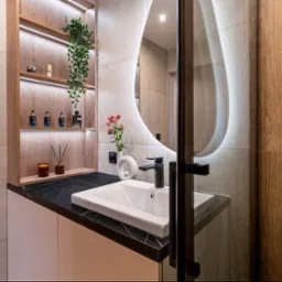 Nowoczesna łazienka z owalnym podświetlanym lustrem, czarnym blatem z marmuru, białą umywalką i drewnianą szafką z półkami podświetlanymi LED, na których stoją flakoniki perfum.