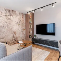 Nowoczesny salon z zabudową regałową z drewna, dekoracyjną tapetą w liście i marmurową szafką RTV pod telewizorem.