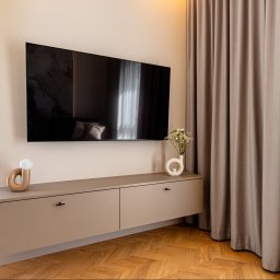 hm-meble marcin heinrich - Nowoczesna, podwieszana szafka RTV w odcieniu taupe z dwoma szufladami, pod telewizorem na ścianie. Na blacie designerska lampa i wazon z kwiatami. Obok beżowe zasłony i drewniana podłoga w jodełkę.