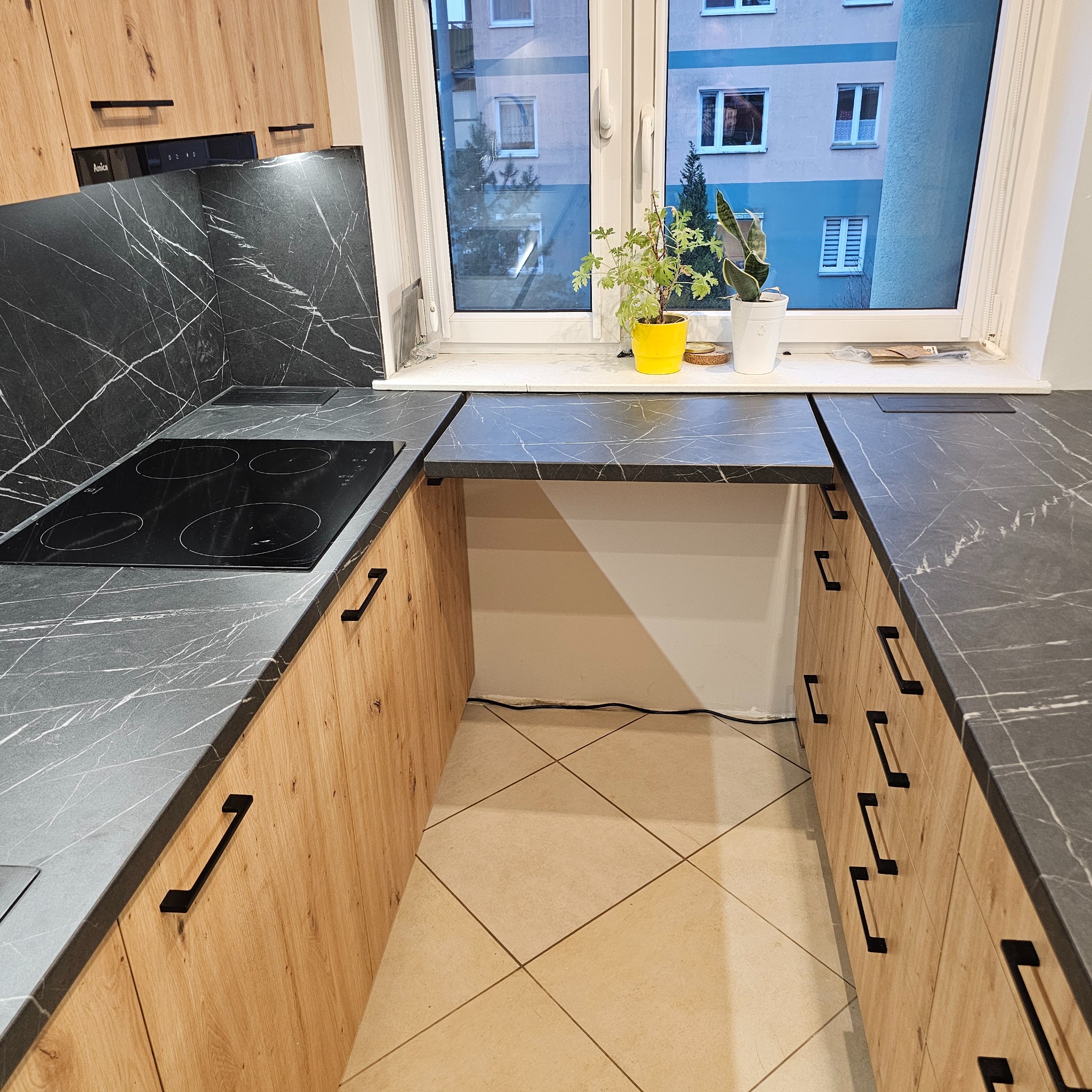 Nowoczesna kuchnia w kształcie litery U z drewnianymi frontami szafek, blatem imitującym marmur i czarnymi uchwytami, z płytą indukcyjną i roślinami doniczkowymi na parapecie okna.