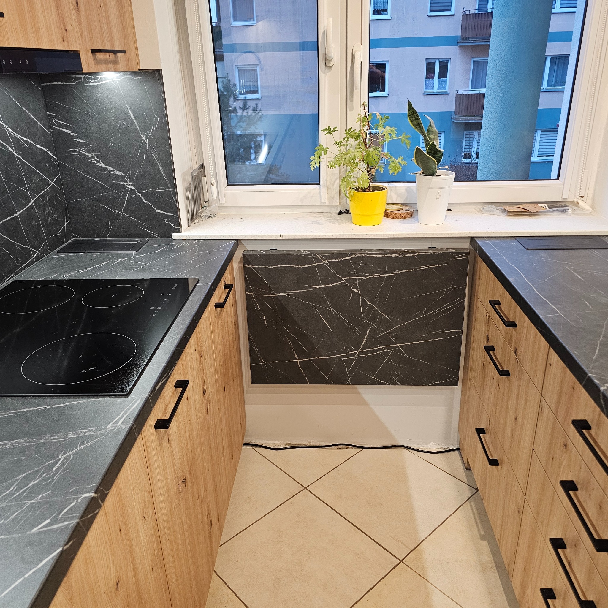 Nowoczesna kuchnia z drewnianymi szafkami i blatem z czarnego marmuru, płyta indukcyjna, widok na okno z roślinami doniczkowymi.