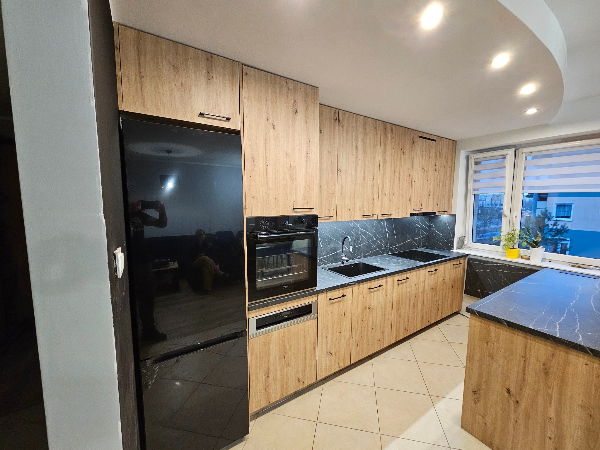 Nowoczesna kuchnia na wymiar z drewnianymi frontami, czarnym blatem i czarną lodówką, widoczne odbicie osoby robiącej zdjęcie w lodówce.