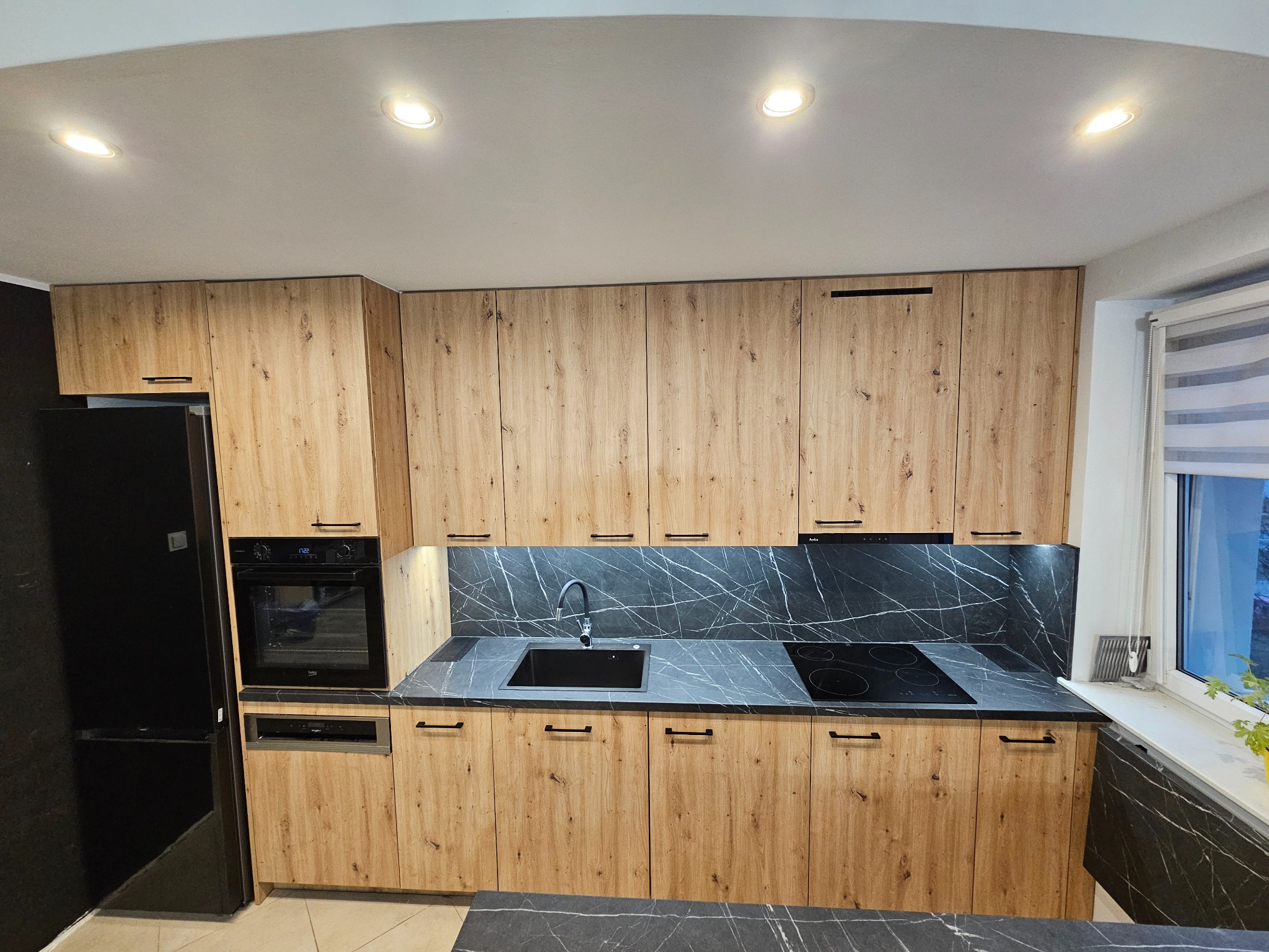 Kompletna zabudowa kuchenna z drewnopodobnymi frontami, czarnym blatem z marmurkowym wzorem i czarnym sprzętem AGD, obejmująca szafki górne i dolne, piekarnik, płytę grzewczą i zlew, oświetlona...