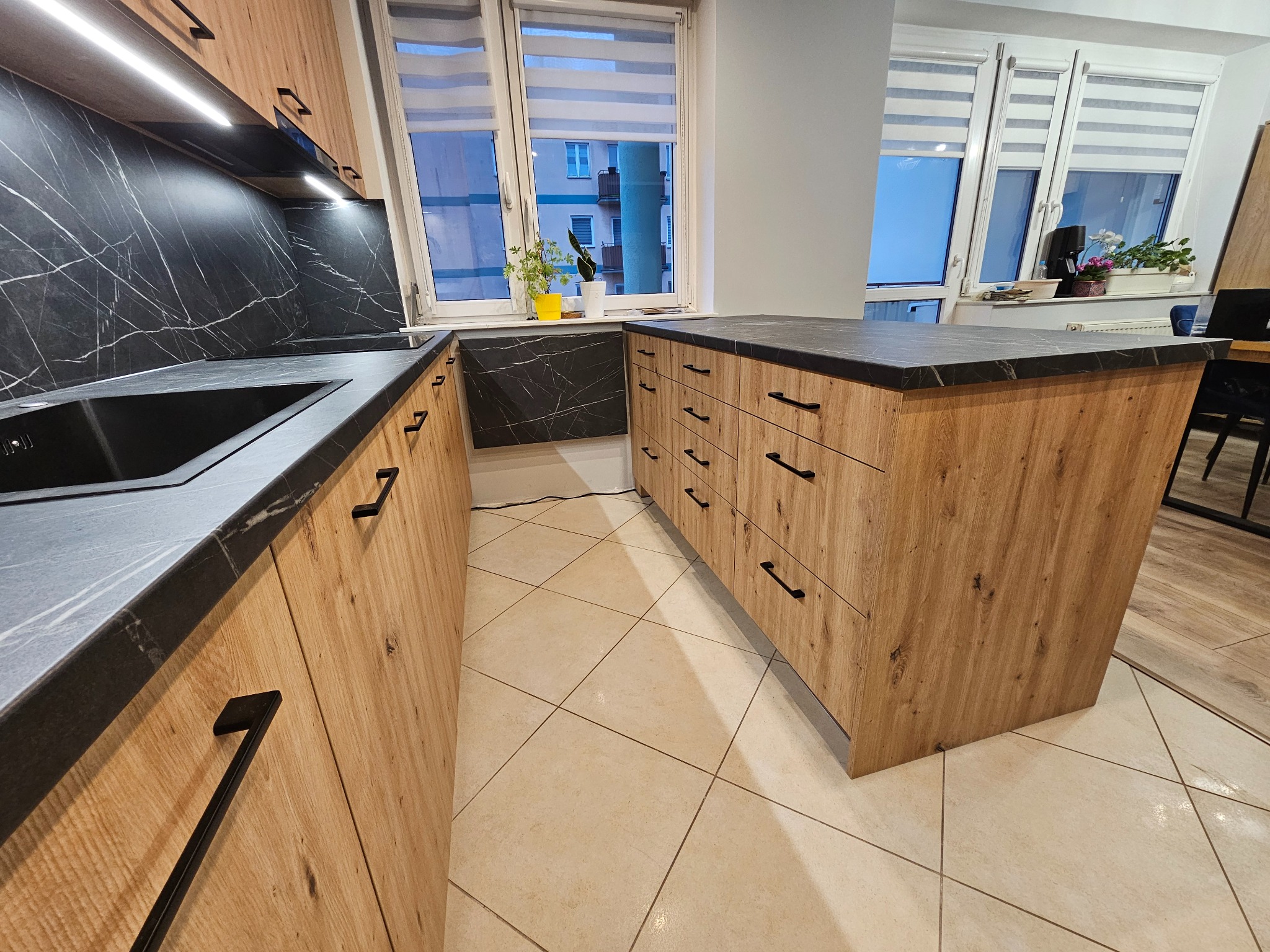 Nowoczesna kuchnia z drewnianymi frontami szafek, czarnym blatem i marmurowym panelem nad zlewem, wyspa kuchenna z szufladami, widok na okno z roletami.