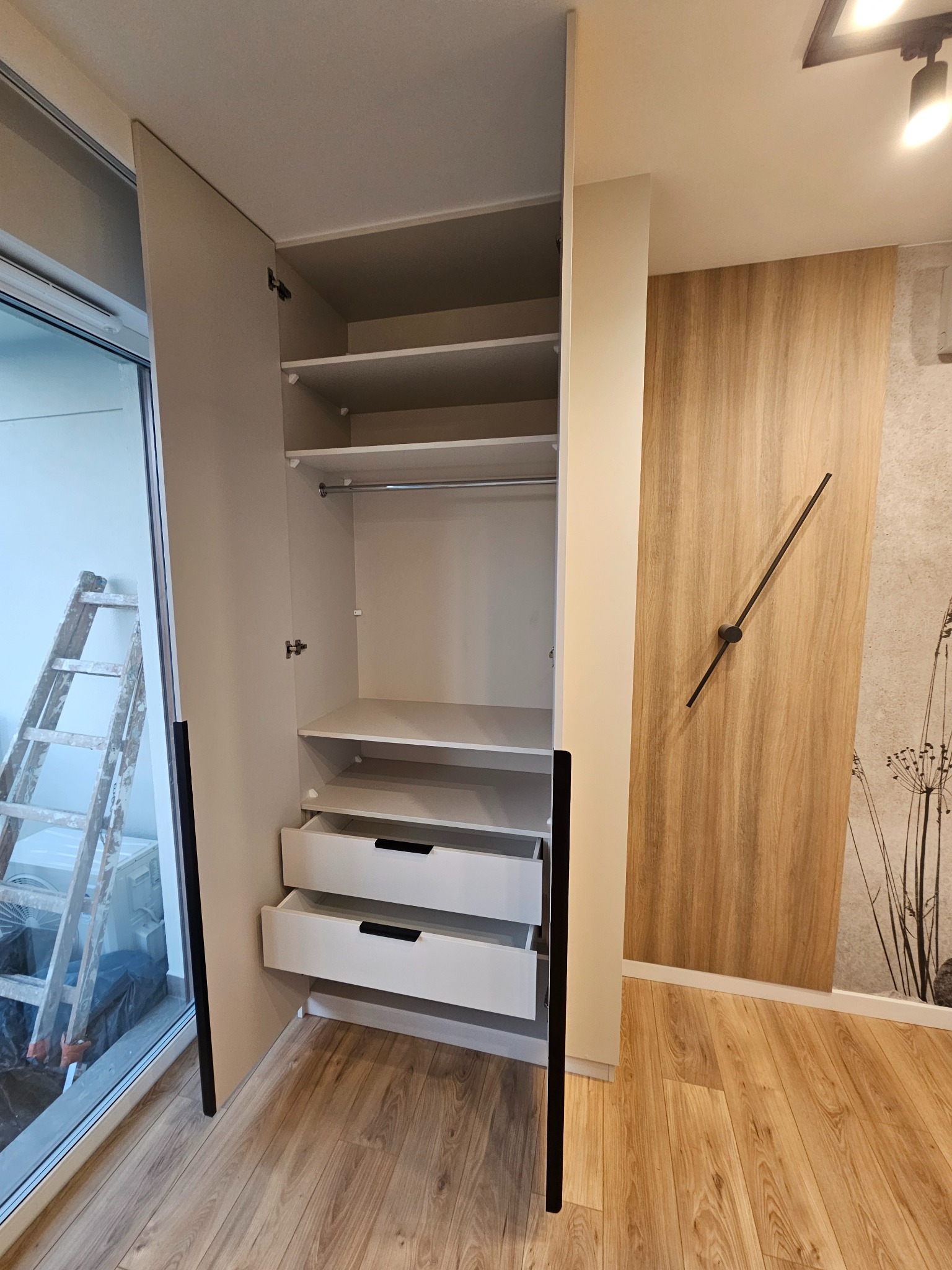 Jasna szafa z otwartymi drzwiami i wysuniętymi szufladami, widoczna drabina oparta o okno balkonowe, drewniana okładzina ścienna z minimalistycznym zegarem.