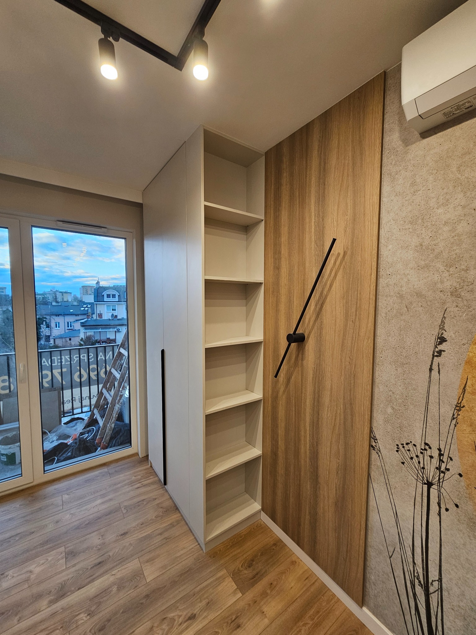 Bydgoszcz: Biała szafa wnękowa z półkami sąsiadująca z drewnianą ścianą z minimalistycznym zegarem, widok z okna na miasto.