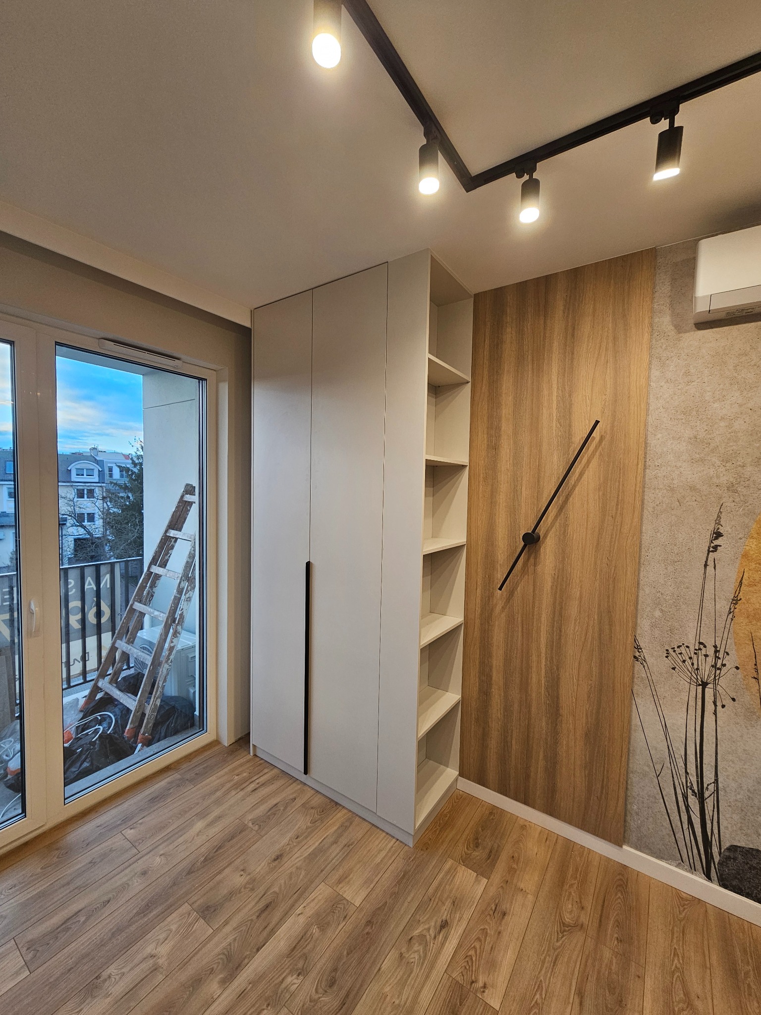 Jasnoszara szafa wnękowa z czarnymi uchwytami, z otwartymi półkami, na podłodze panele, na ścianie dekoracyjny panel drewnopodobny z zegarem, widok na balkon z drabiną