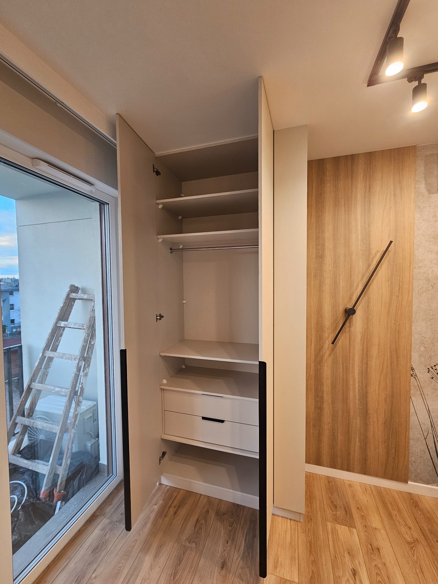 Otwarta, jasna szafa wnękowa z półkami i szufladą, widoczna drabina oparta o okno balkonowe, drewniana okładzina ściany z minimalistycznym zegarem.