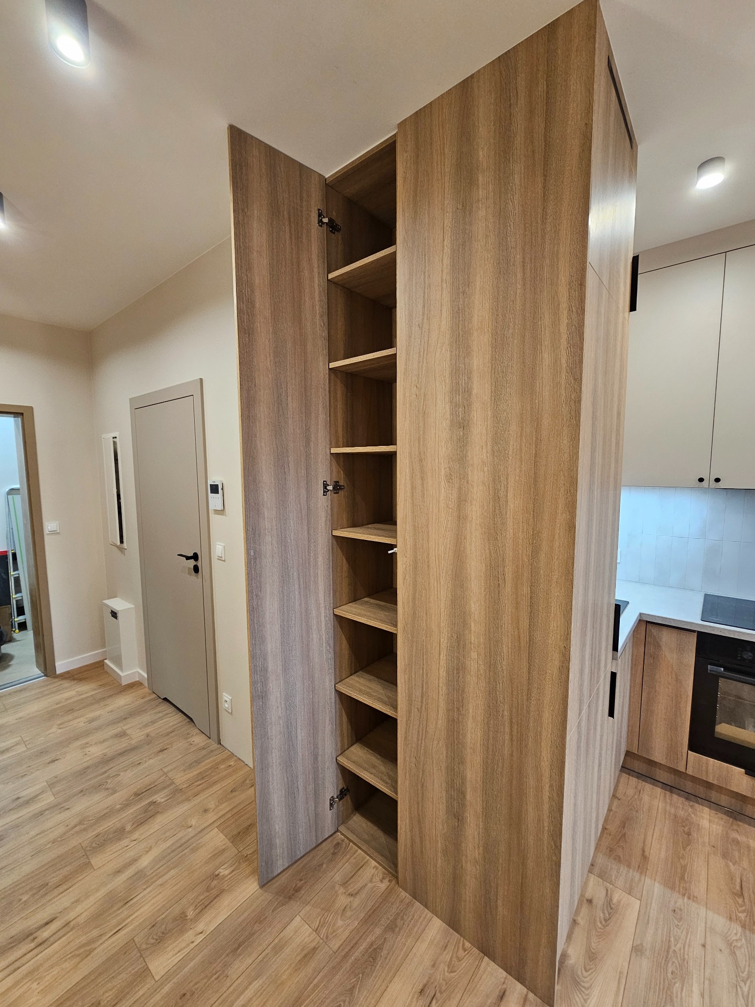 Wysoka, drewniana szafa wnękowa z otwartymi drzwiami, ukazująca puste półki, wkomponowana w przestrzeń między przedpokojem a kuchnią, z podłogą imitującą drewno.
