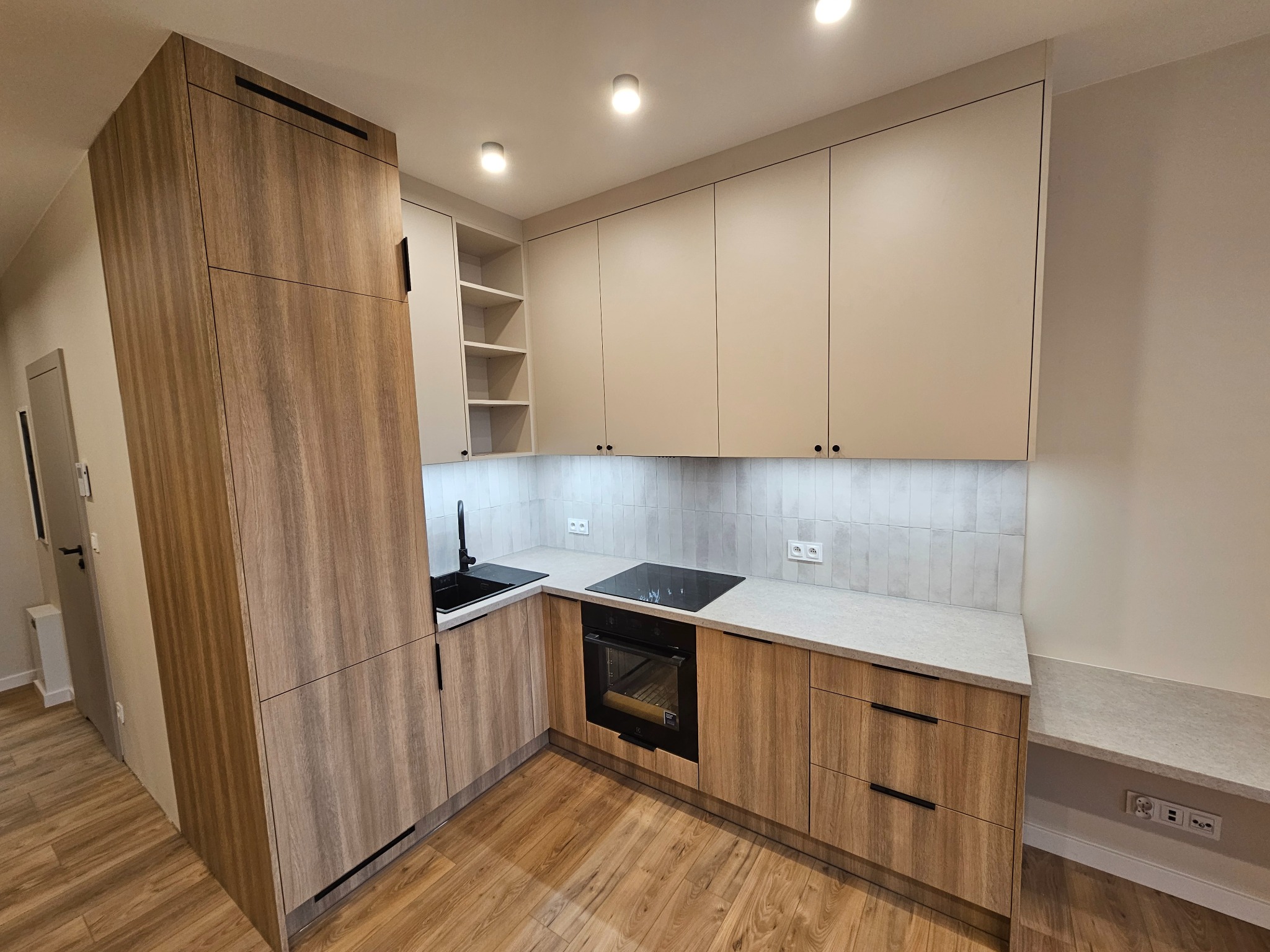 Nowoczesna kuchnia z frontami w odcieniach beżu i drewna, czarnym zlewem i płytą indukcyjną, oświetlona punktowymi lampami sufitowymi, z widocznymi gniazdkami elektrycznymi nad blatem.