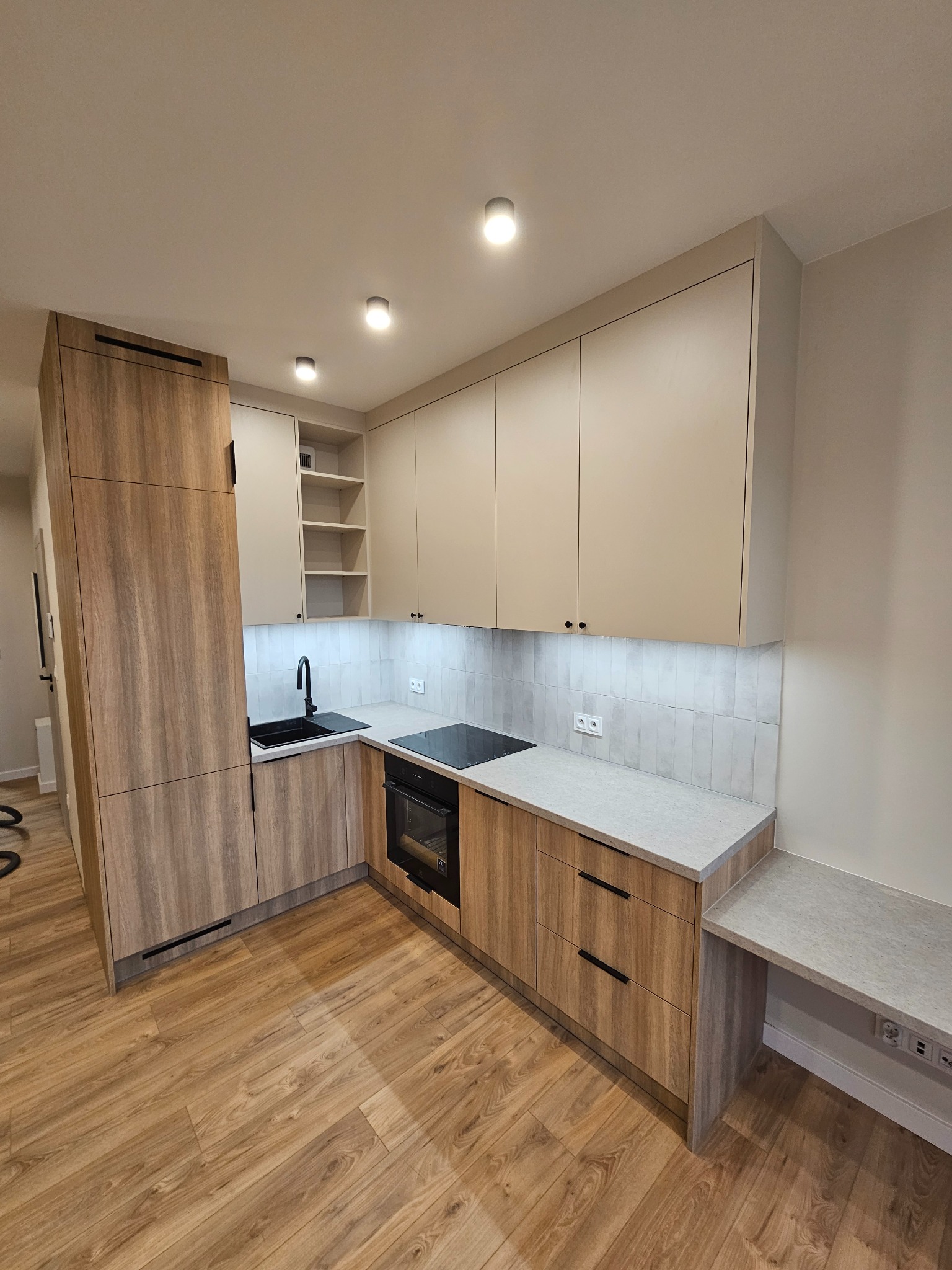 Nowoczesna kuchnia z drewnianymi frontami szafek dolnych i jasnymi górnymi, czarny zlew i płyta indukcyjna, szara ściana nad blatem z podłużnych płytek, oświetlenie podszafkowe LED.