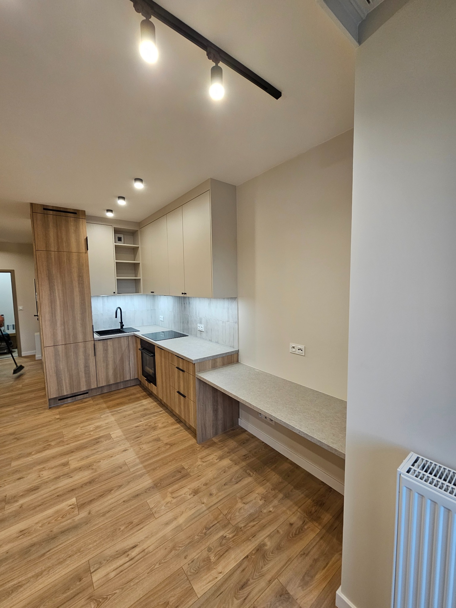 Nowoczesna kuchnia z zabudową w odcieniach beżu i drewna, zintegrowana z blatem roboczym pełniącym funkcję biurka, oświetlona listwą LED pod szafkami i reflektorami sufitowymi.