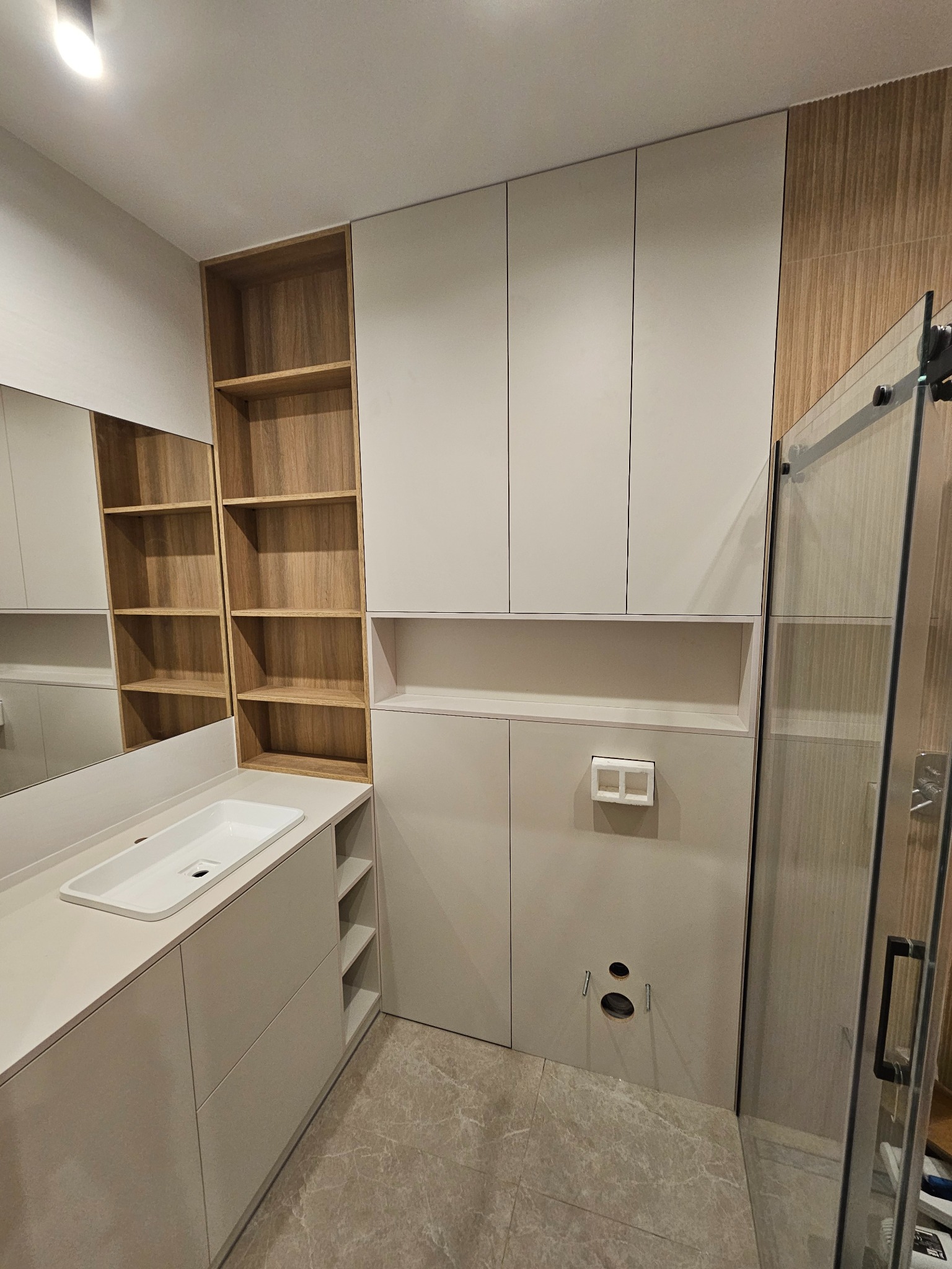 Nowoczesna łazienka z zabudową meblową: białe szafki wiszące i stojące, drewniane półki otwarte, umywalka nablatowa, szklana kabina prysznicowa i płytki podłogowe imitujące marmur. Widoczne otwory...