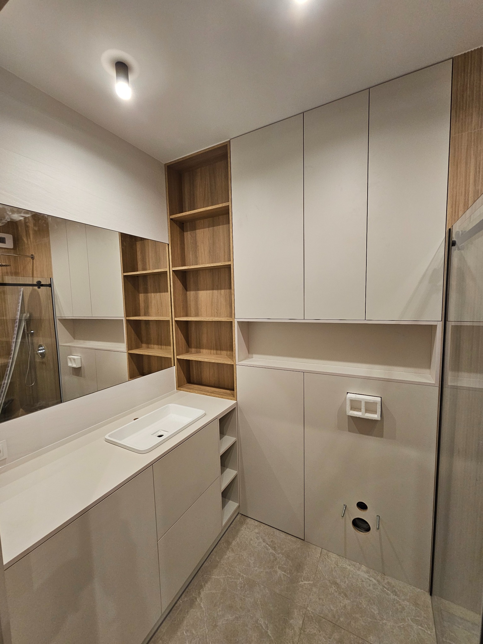 Nowoczesna łazienka z zabudową meblową na wymiar w odcieniach bieli i drewna; widoczna umywalka wpuszczana w blat, szafki wiszące i regał otwarty z drewnianymi półkami.