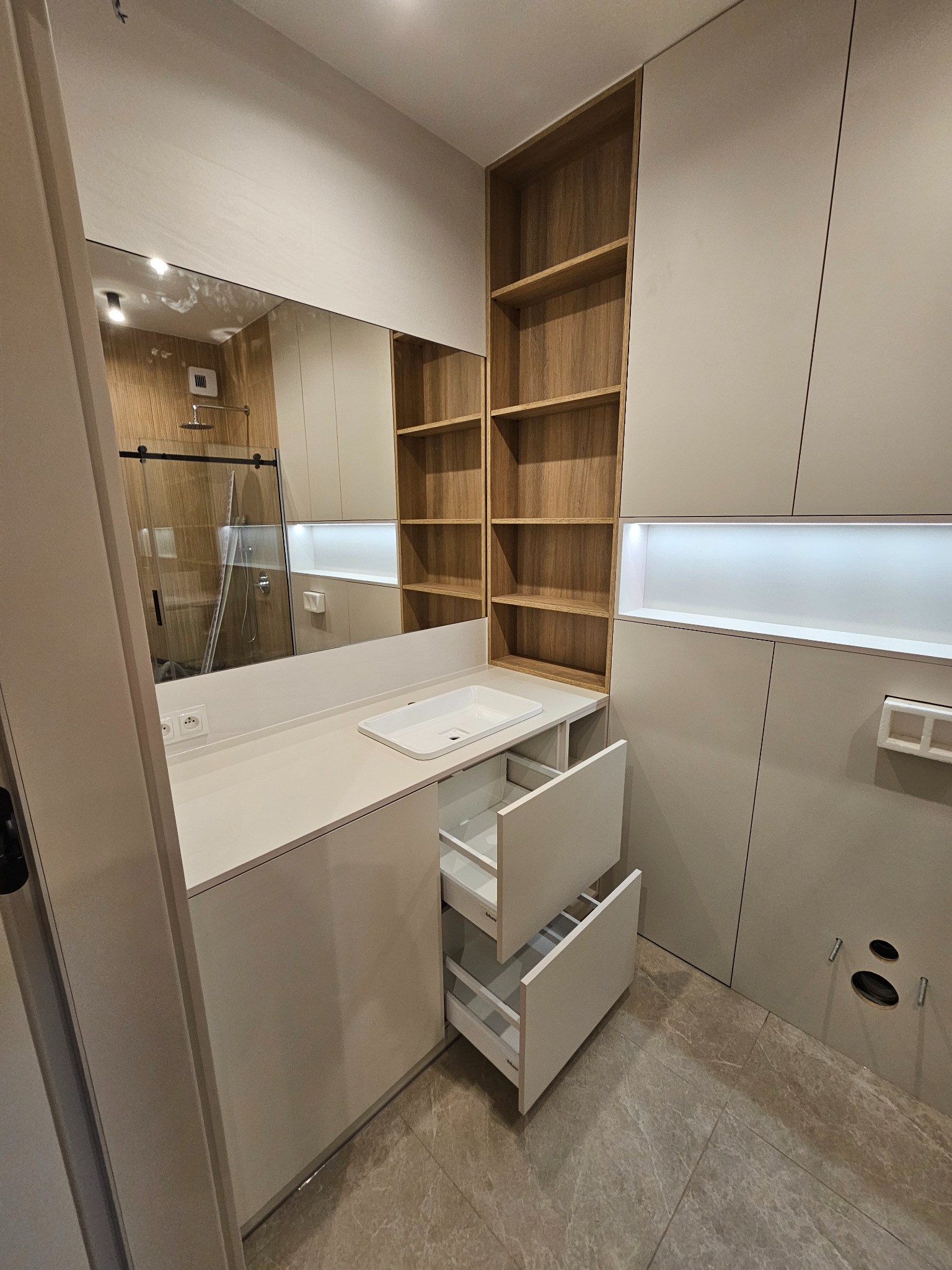 Nowoczesna łazienka z zabudową meblową w odcieniach szarości i drewna, z otwartymi szufladami pod umywalką i półkami na ścianie. W lustrze odbija się kabina prysznicowa.