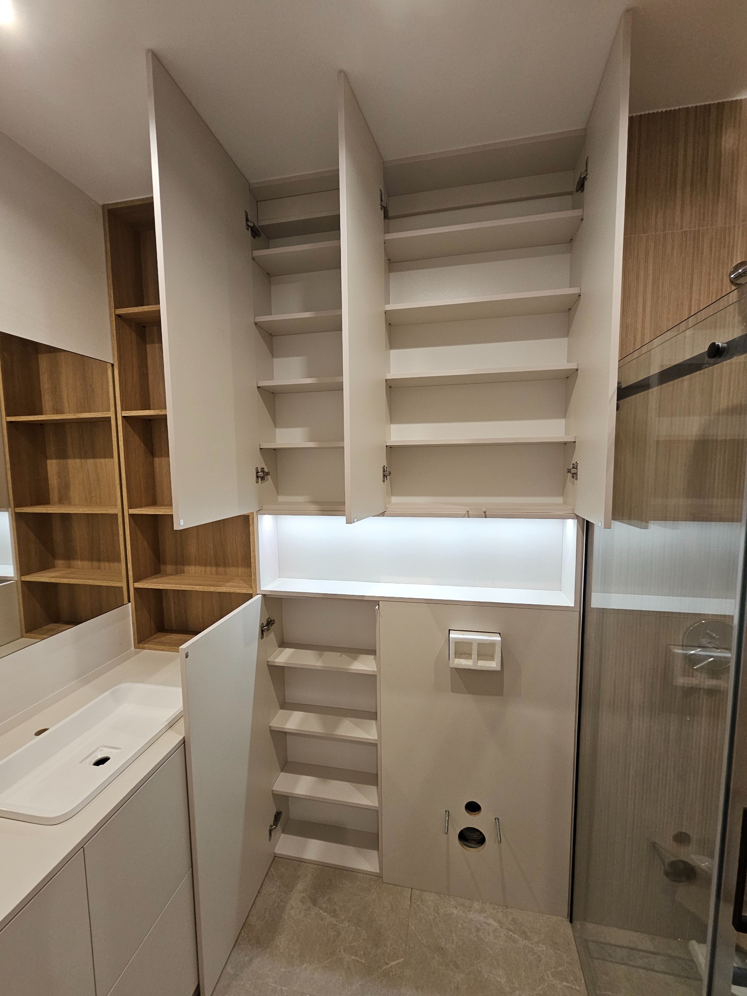 Nowoczesna łazienka z szafkami z białymi frontami i drewnianymi półkami, otwarte drzwi ukazują półki, widoczny zlew i kabina prysznicowa z przezroczystego szkła.