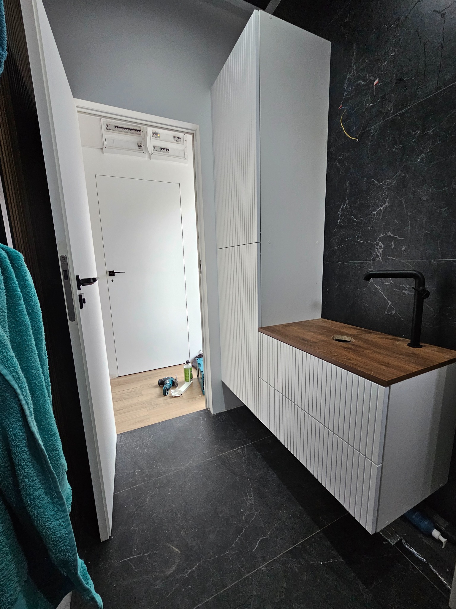 Biała szafka łazienkowa na wymiar z drewnianym blatem i czarną armaturą na tle ściany z czarnego marmuru, widoczne narzędzia w tle wskazują na trwający montaż.