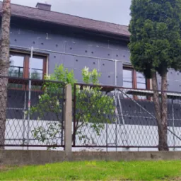 Dom w trakcie ocieplania elewacji, widoczne szare płyty izolacyjne z charakterystycznymi otworami, rusztowanie, brązowe ramy okienne, fragment ogrodu z kwitnącym krzewem bzu i tujami, ujęcie...