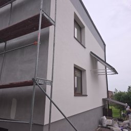 FACH-BUD - Dwukondygnacyjny dom z rusztowaniem w trakcie prac elewacyjnych; dolna część w szarym tynku, górna w jasnym. Widoczne okna i zadaszenie nad wejściem.