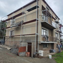 FACH-BUD - Dwupiętrowy dom w trakcie prac elewacyjnych, otoczony rusztowaniem. Widoczni pracownicy, materiały budowlane i fragment posesji z kostki brukowej.