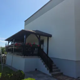 Dwukondygnacyjny dom z werandą i schodami wejściowymi, świeżo otynkowany na biało-szaro, z ciemnym drewnem werandy i czerwonymi kwiatami na balkonie.