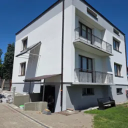 Dwukondygnacyjny budynek z balkonami, świeżo po malowaniu elewacji na biało i szaro, z widocznym fragmentem prac przy wejściu i stosem materiałów budowlanych.