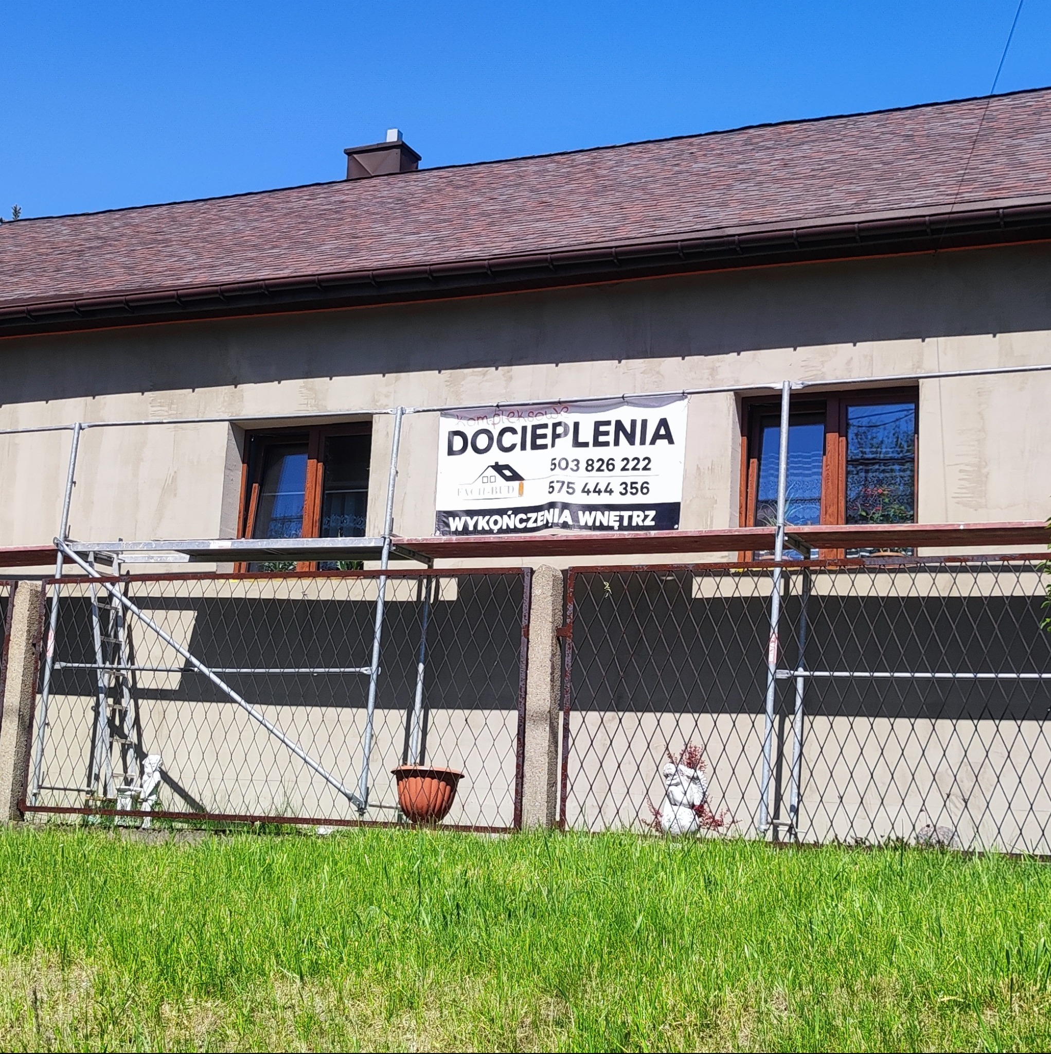 Dom jednorodzinny z szarą elewacją w trakcie prac, widoczne rusztowanie i baner reklamowy firmy, brązowy dach z dachówki bitumicznej.