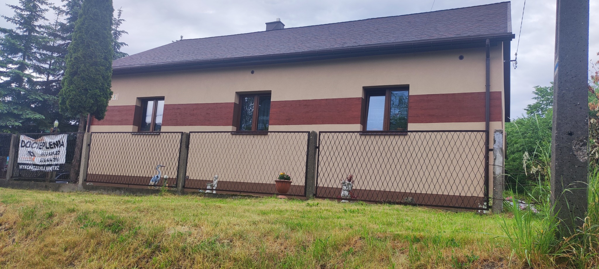 Dwukondygnacyjny dom z beżową elewacją i brązowym pasem dekoracyjnym, widoczny zza ażurowego ogrodzenia z figurkami ogrodowymi i donicą z kwiatami.