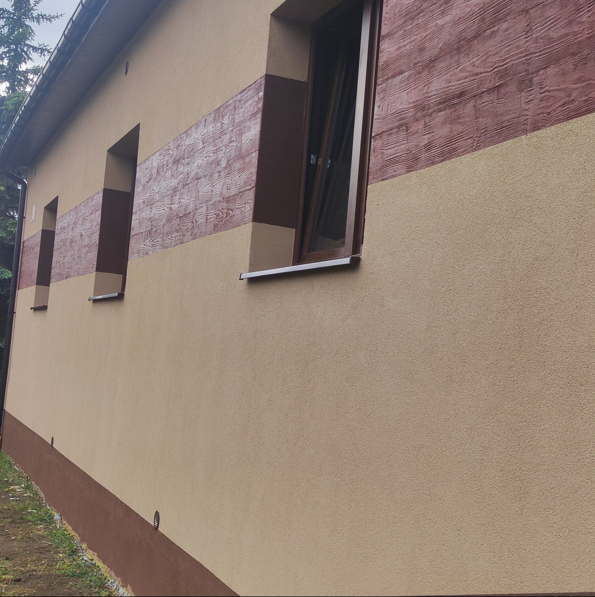 Elewacja domu z beżowym tynkiem i dekoracyjnym pasem z fakturą drewna w odcieniach brązu, widoczne okna z brązowymi ramami i parapetami.