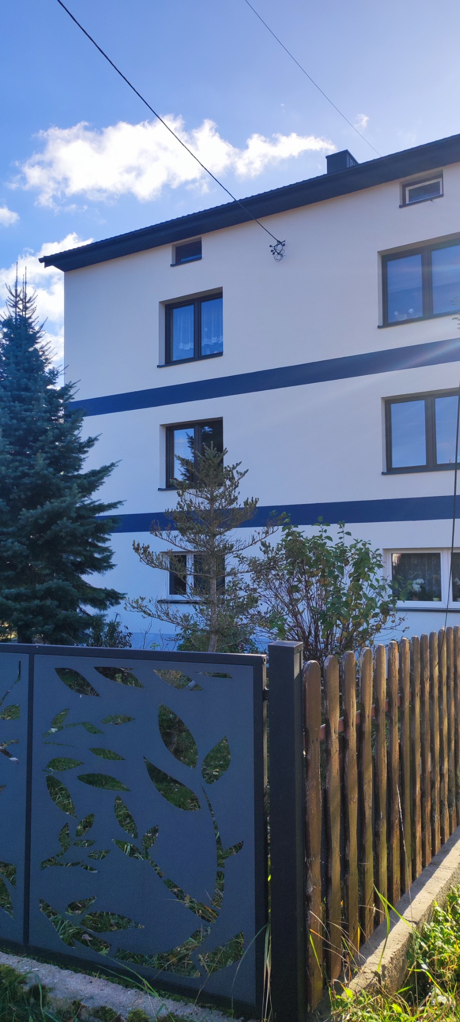 Dwupiętrowy dom z białą elewacją i granatowymi pasami, widoczny zza ozdobnego metalowego i drewnianego ogrodzenia, drzewa iglaste przed budynkiem.