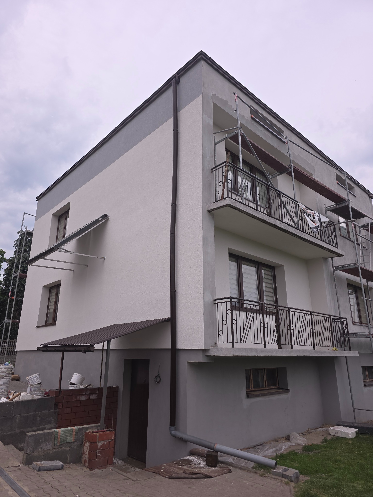 Dwukolorowa elewacja budynku w Zawierciu, częściowo otoczona rusztowaniem. Widoczne balkony z metalowymi balustradami i okna z roletami. Budynek po remoncie.