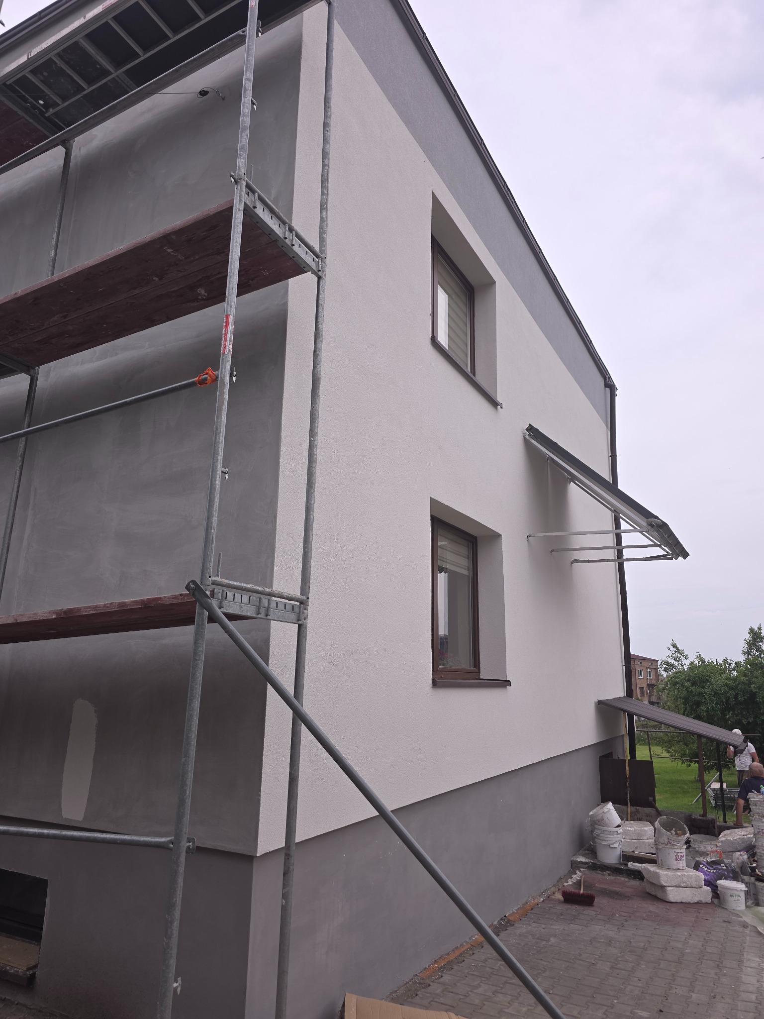 Dwukondygnacyjny dom z rusztowaniem w trakcie prac elewacyjnych; dolna część w szarym tynku, górna w jasnym. Widoczne okna i zadaszenie nad wejściem.