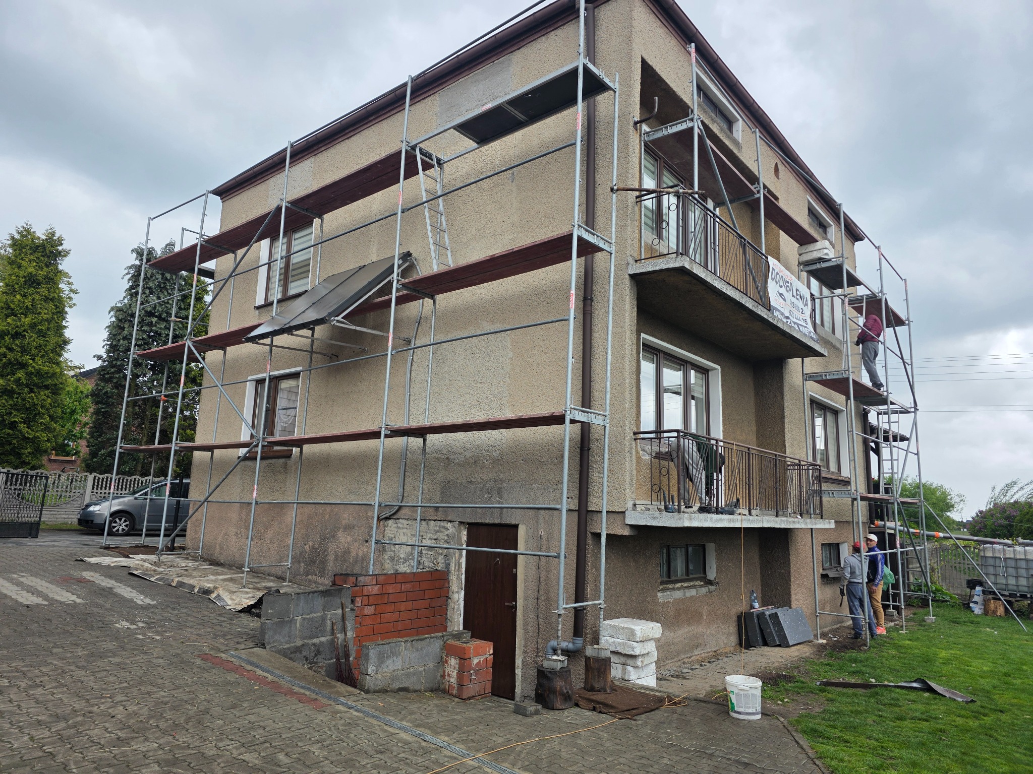Dwupiętrowy dom w trakcie prac elewacyjnych, otoczony rusztowaniem. Widoczni pracownicy, materiały budowlane i fragment posesji z kostki brukowej.