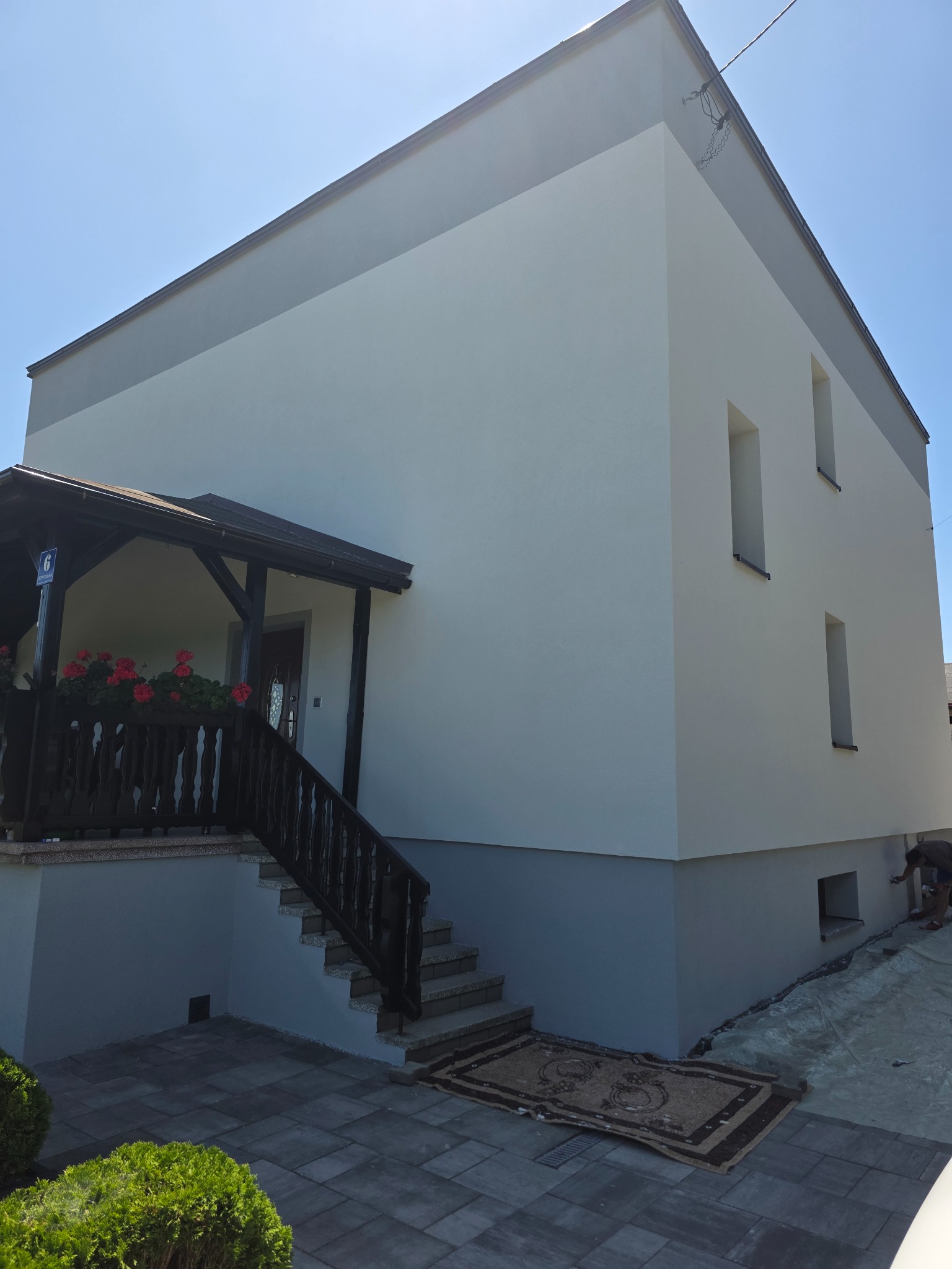 Dwukondygnacyjny dom z nową, dwukolorową elewacją (biało-szarą). Widoczne schody wejściowe z balustradą i kwiatami oraz fragment pracującego człowieka przy elewacji.