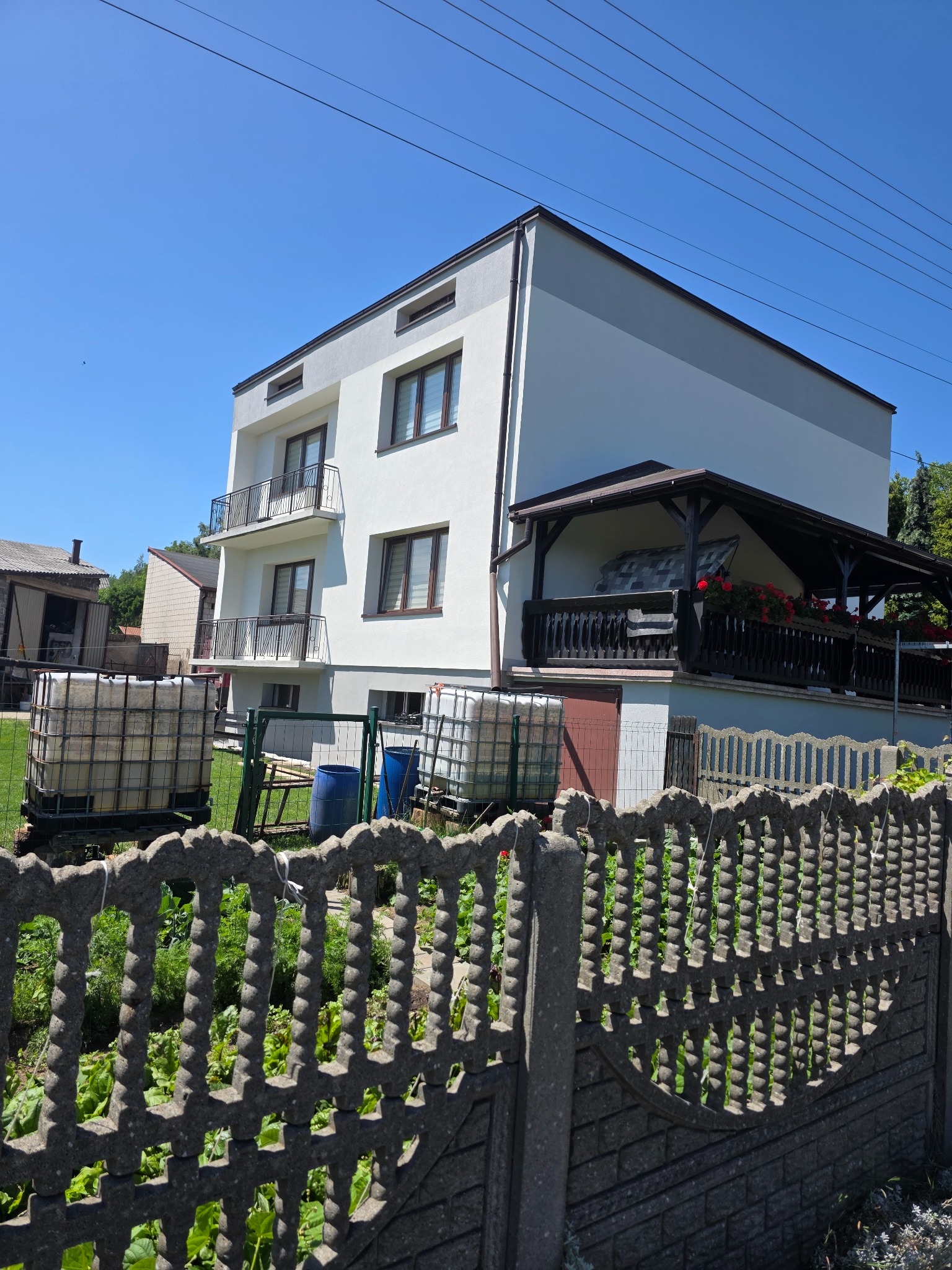 Dwukondygnacyjny dom z balkonami i zadaszonym tarasem, widoczny zza ozdobnego betonowego ogrodzenia i zbiorników na wodę, w słoneczny dzień.
