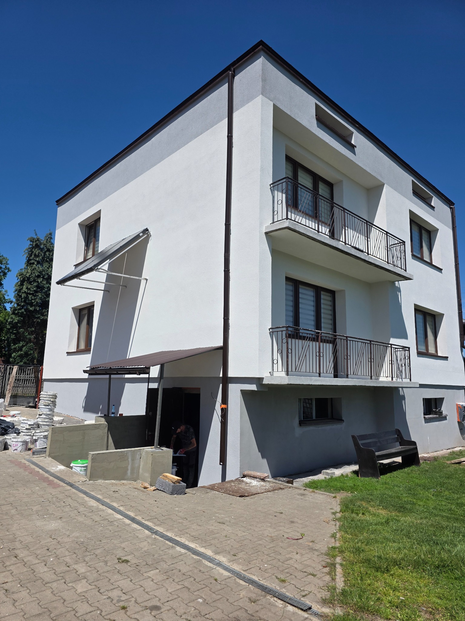 Dwukondygnacyjny budynek z balkonami, świeżo po malowaniu elewacji na biało i szaro, z widocznym fragmentem prac przy wejściu i stosem materiałów budowlanych.